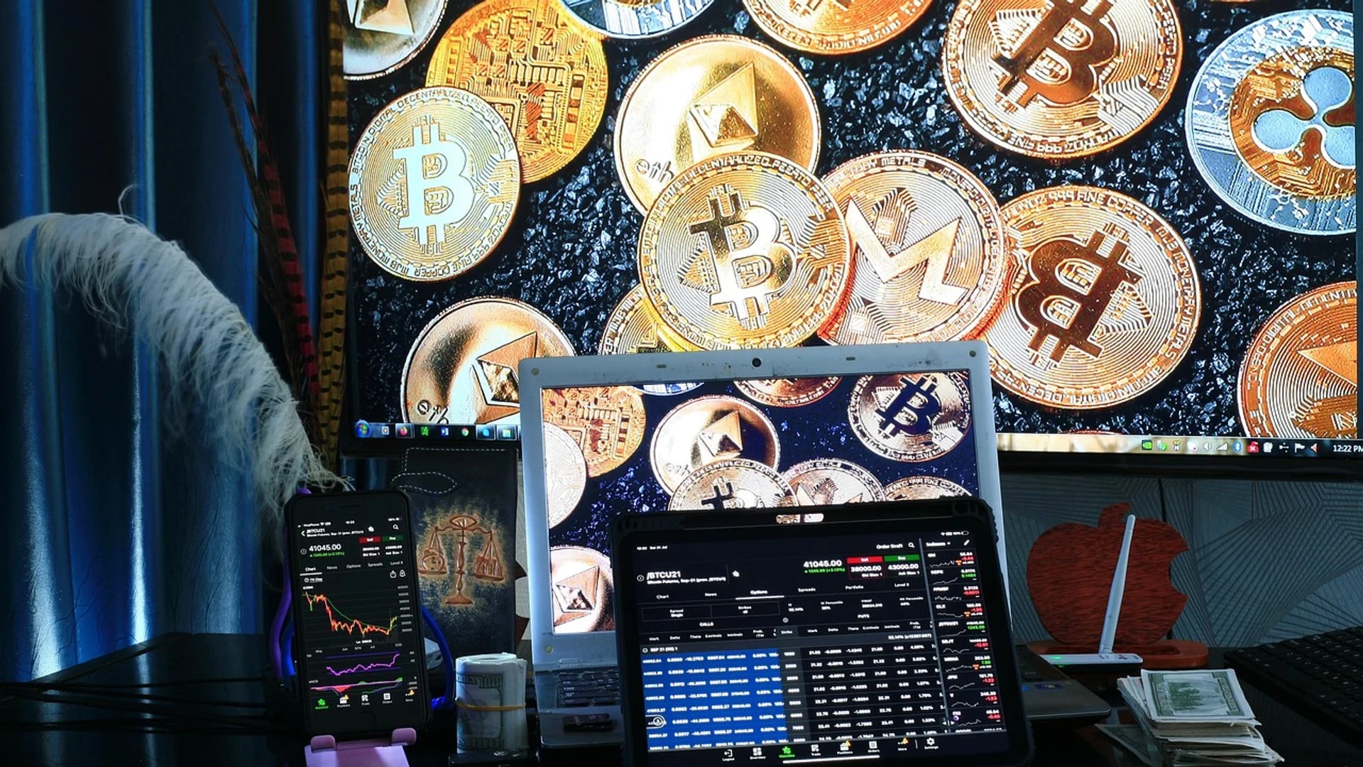 A desk with a laptop, tablet, and smartphone displaying cryptocurrency trading charts in front of a monitor showing gold cryp